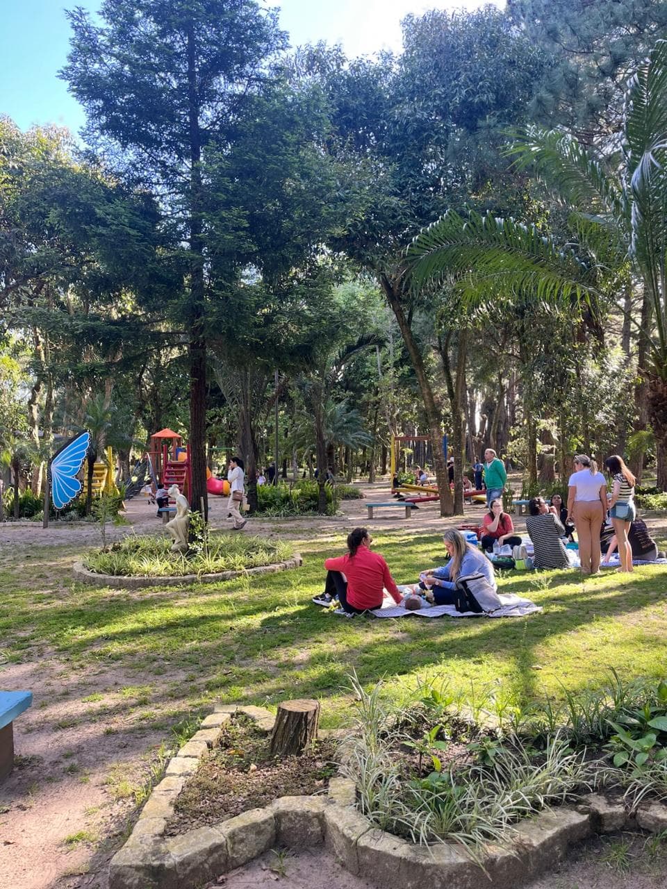Fotografia de um parque arborizado num dia ensolarado, com várias pessoas a desfrutar do espaço ao ar livre. Em primeiro plano e ao centro, grupos de pessoas fazem um piquenique, sentadas sobre toalha