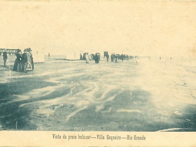 Fotografia histórica, com tons sépia e um ligeiro azul esbatido, que retrata uma ampla extensão de praia. Em primeiro plano, a areia exibe padrões ondulados marcados pela água ou pelo vento. Ao fundo,