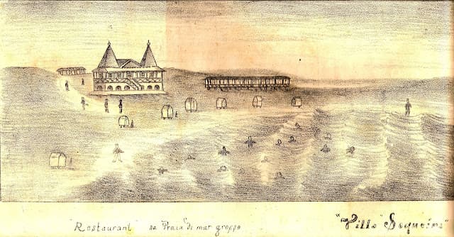 Fotografia histórica, com tons sépia e um ligeiro azul esbatido, que retrata uma ampla extensão de praia. Em primeiro plano, a areia exibe padrões ondulados marcados pela água ou pelo vento. Ao fundo, espalhados ao longo do areal, encontram-se grupos de pessoas vestidas com trajes formais de época, incluindo vestidos compridos, casacos e chapéus. Perto das pessoas, veem-se algumas estruturas retangulares brancas, que se assemelham a antigas cabines de banho. Na margem inferior da imagem, está impressa a legenda: "Vista da praia balnear — Villa Sequeira — Rio Grande".